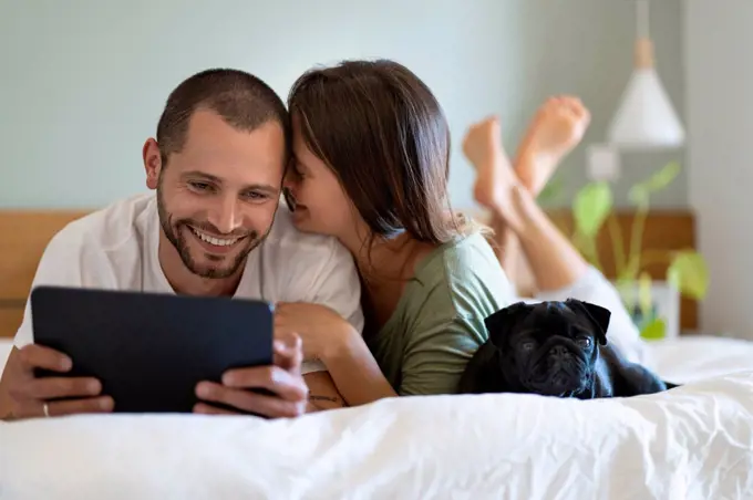 Woman embracing boyfriend looking at digital tablet while lying on bed in bedroom at home