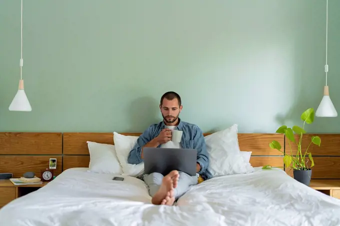 Young man having coffee using laptop while sitting on bed at home