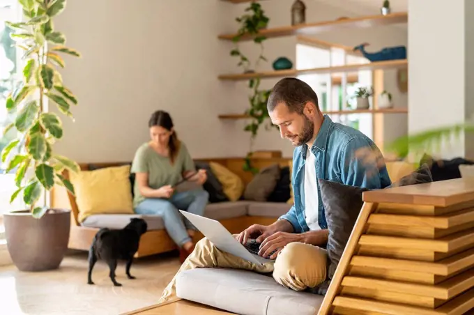 Young man working on laptop while wife looking at Pug dog in background at home