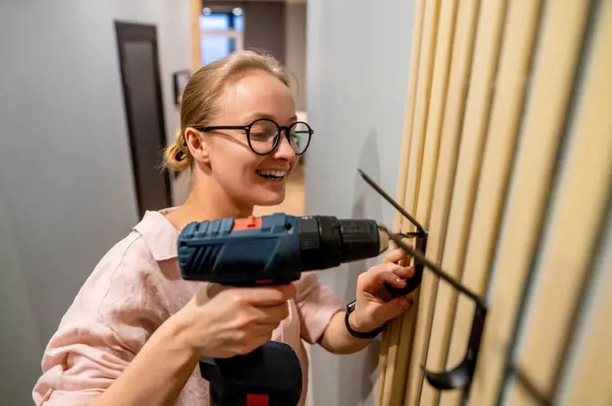 Smiling woman using drill machine while fixing hook on wall at home