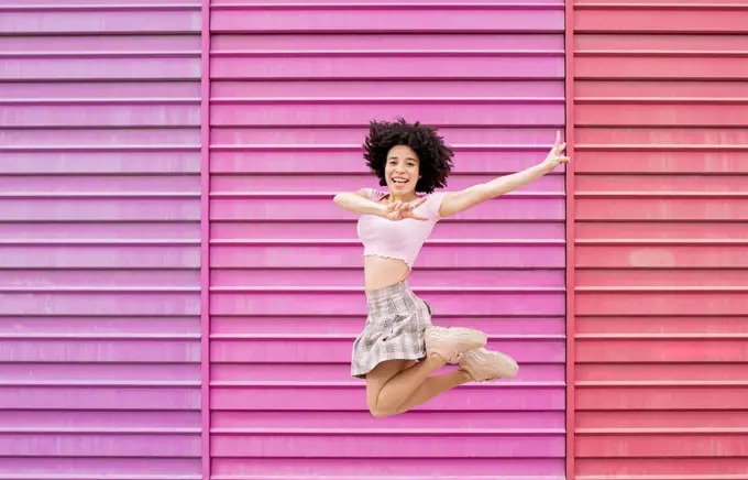 Carefree woman showing peace sign while jumping against multi colored wall