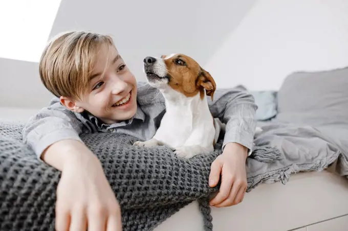 Smiling boy with arm around dog lying on bed at home