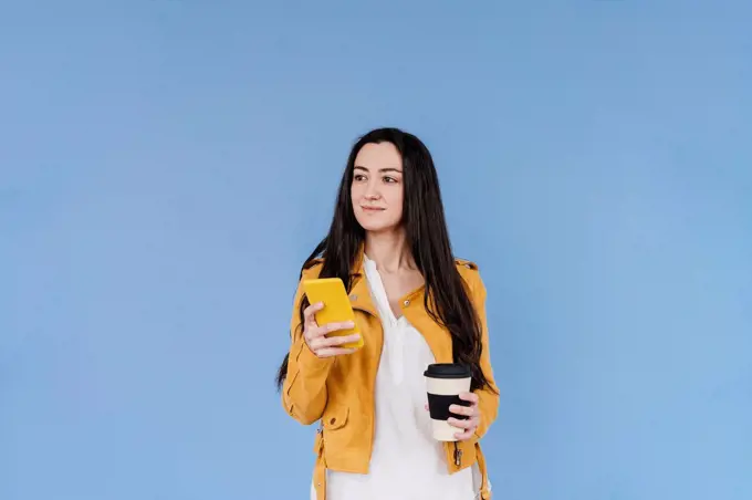 Smiling woman with mobile phone looking away while standing against blue background