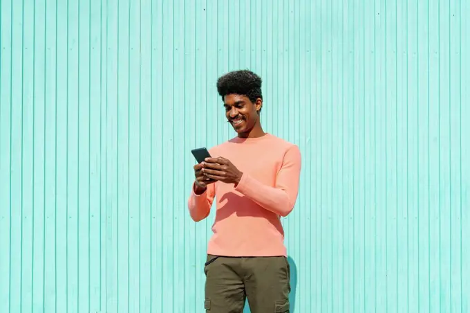 Smiling man using smart phone in front of turquoise wall during sunny day