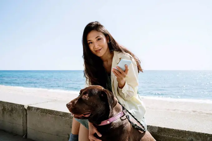 Smiling woman sitting with Labrador Retriever by sea