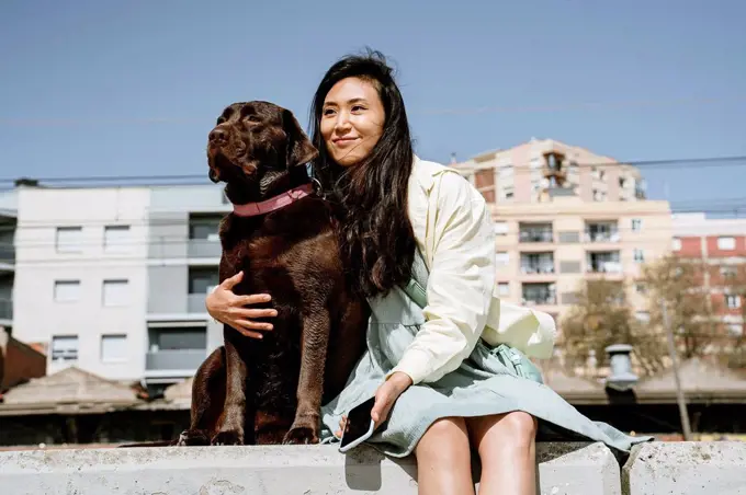 Smiling woman sitting with Chocolate Labrador on retaining wall in front of building