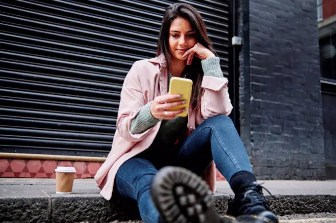 Beautiful woman with coffee cup using mobile phone while sitting on sidewalk