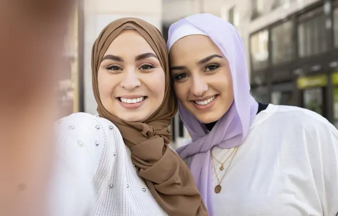 Smiling Arab female friends in hijabs taking selfie in city