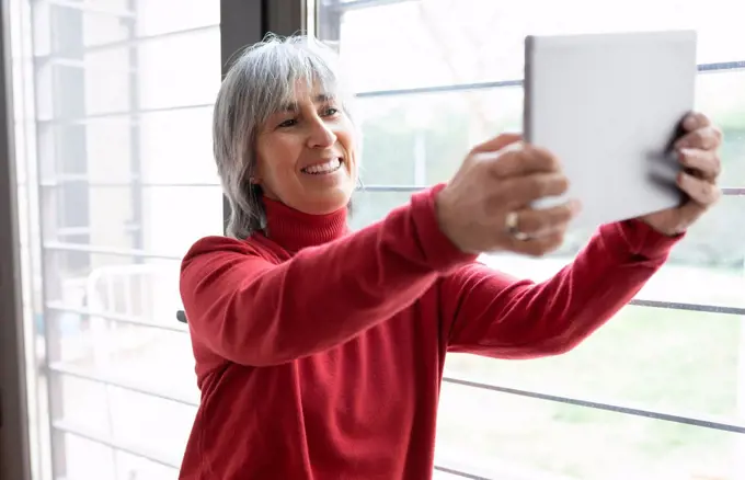 Smiling woman taking selfie through digital tablet by window at home