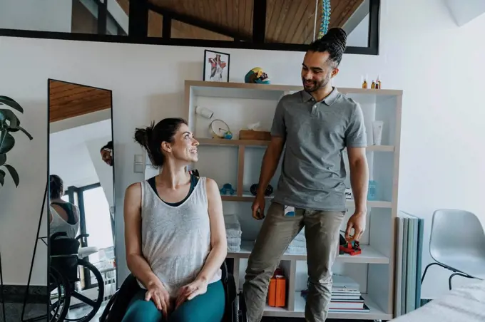 Smiling physiotherapist with female patient sitting on wheelchair in medical practice