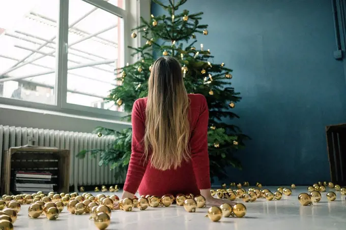 Back view of woman sitting on the floor with many golden Christmas baubles looking at Christmas tree