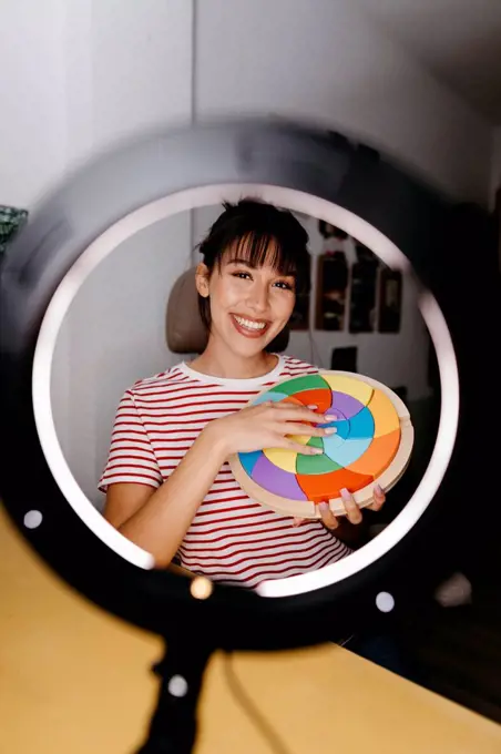 Smiling female influencer seen through ring light while holding multi colored beauty product