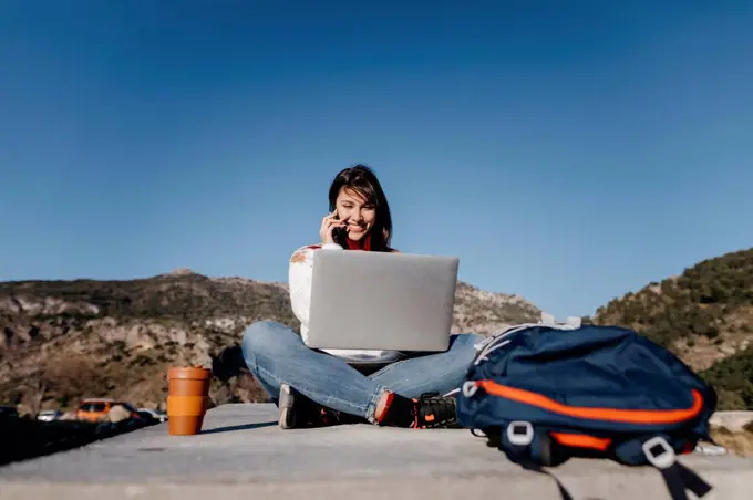 Smiling woman talking on phone while sitting on concrete at mountain