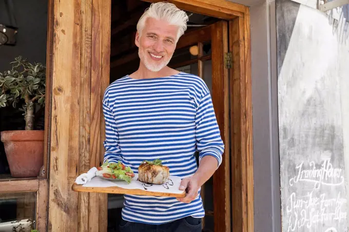 Mature man standing in front of his restaurant, serving a dish