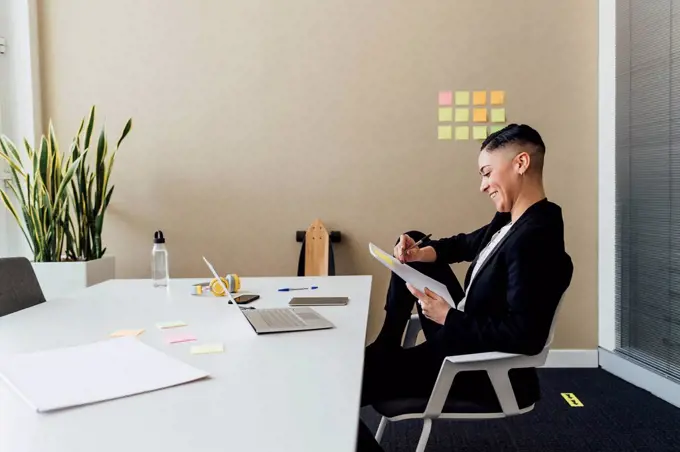 Smiling businesswoman writing on paper while sitting at desk in office