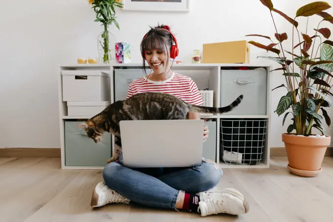 Young woman wearing headphones sitting with laptop and cat at home