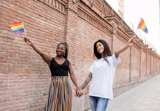 Multi-ethnic lesbian couple holding flag while standing on footpath near wall