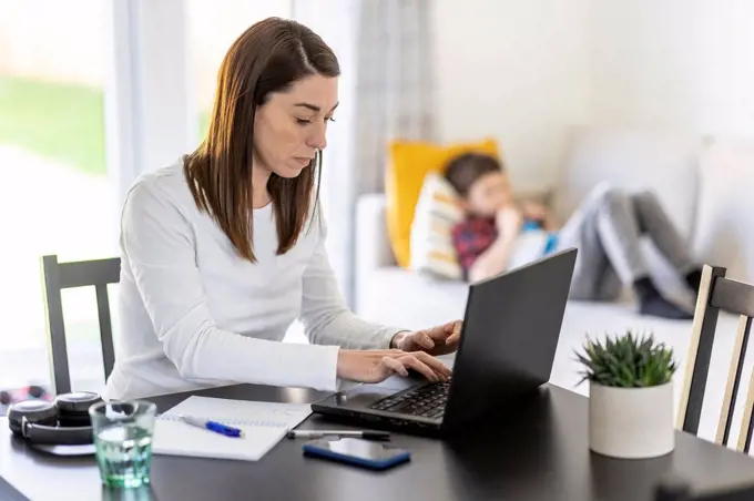 Female entrepreneur working on laptop with son in background at home