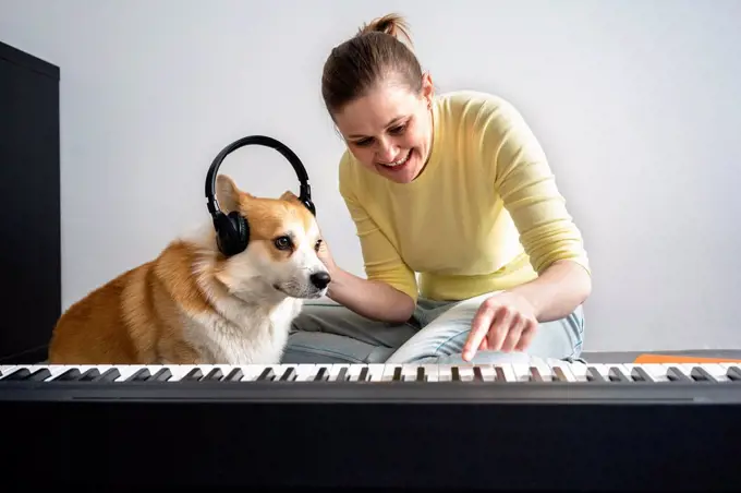 Smiling woman with dog wearing headphones to dog while practicing piano in living room