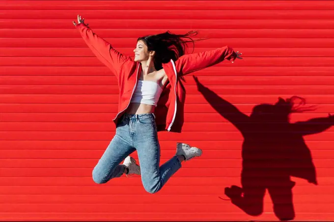 Smiling teenage girl with arms outstretched jumping in front of red wall