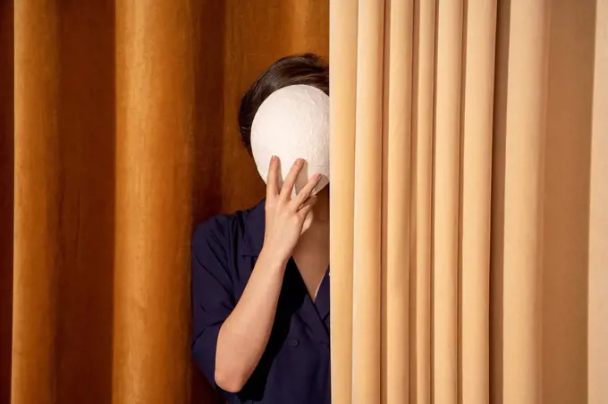 Woman covering face with white mask by curtain