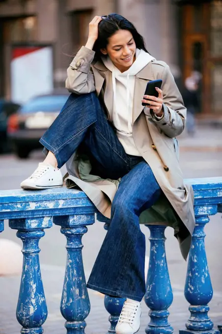 Smiling woman using mobile phone while sitting on blue railing