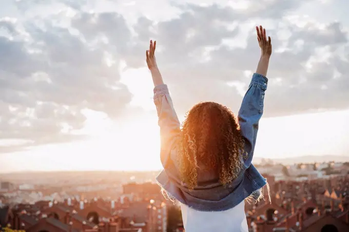 Woman with arms raised looking at city during sunset