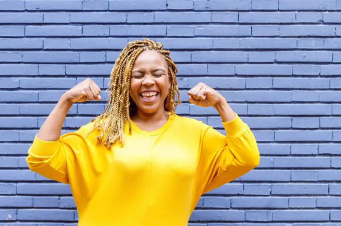 Cheerful plus size woman with eyes closed flexing muscles in front of brick wall