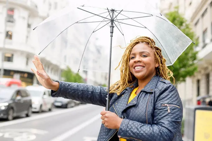 Happy woman with braids holding umbrella while hailing taxi in city