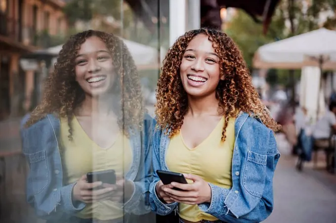 Smiling woman with smart phone leaning on glass wall