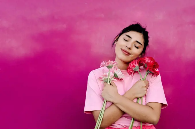 Young woman with arms crossed holding bunch of Gerbera daisies in front of pink wall