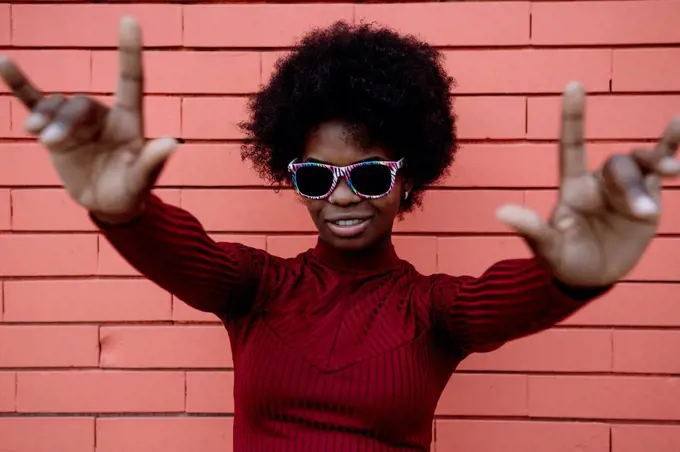 Young woman with sunglasses gesturing near brick wall