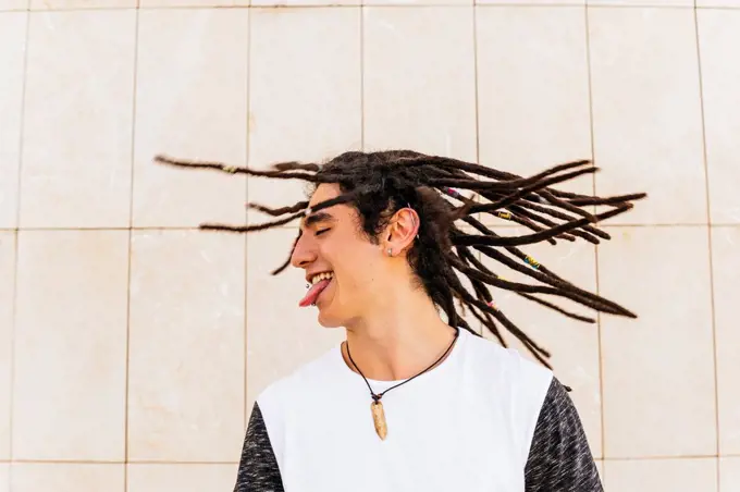 Man with dreadlocks shaking head while sticking out tongue in front of wall