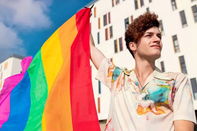 Young man looking away while blowing rainbow flag