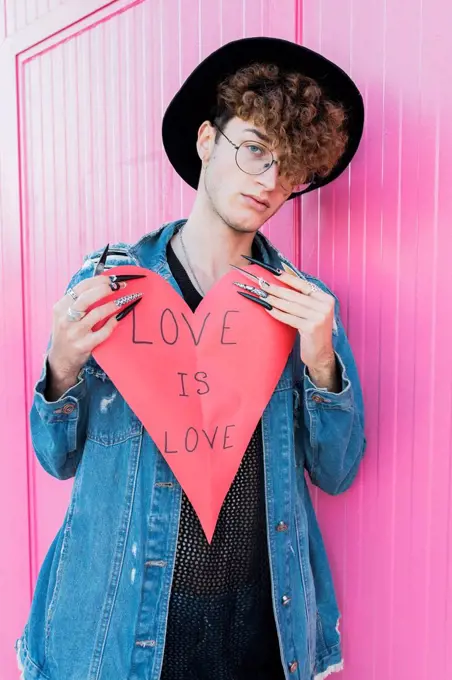 Young gay man with heart shape leaning on pink wall