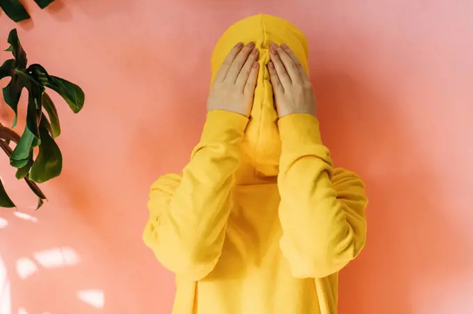 Young woman covering face with yellow hoodie in front of peach wall