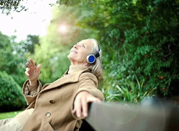 Smiling senior woman looking up while listening music on bench at public park