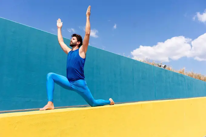 Man with arms raised practicing yoga on retaining wall during sunny day