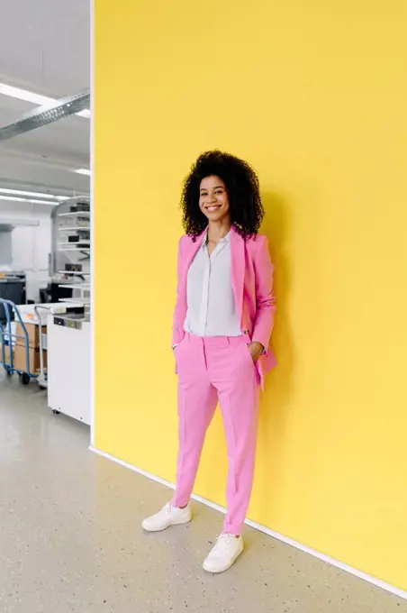 Smiling female entrepreneur with hands in pockets standing in front of yellow wall