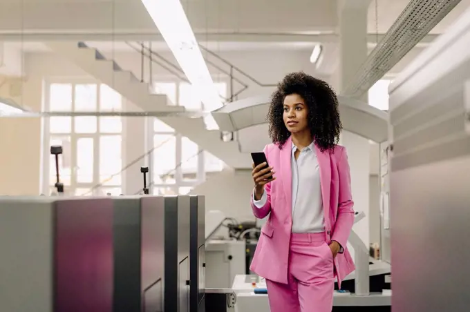 Female professional with mobile phone looking away while standing amidst machines in industry