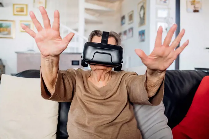 Senior woman wearing virtual reality simulator gesturing while siting on sofa in living room at home