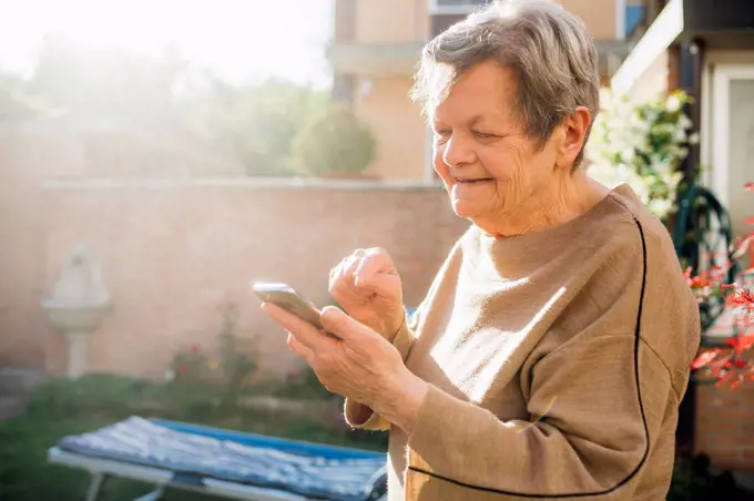 Smiling senior woman using smart phone while standing at backyard during sunny day
