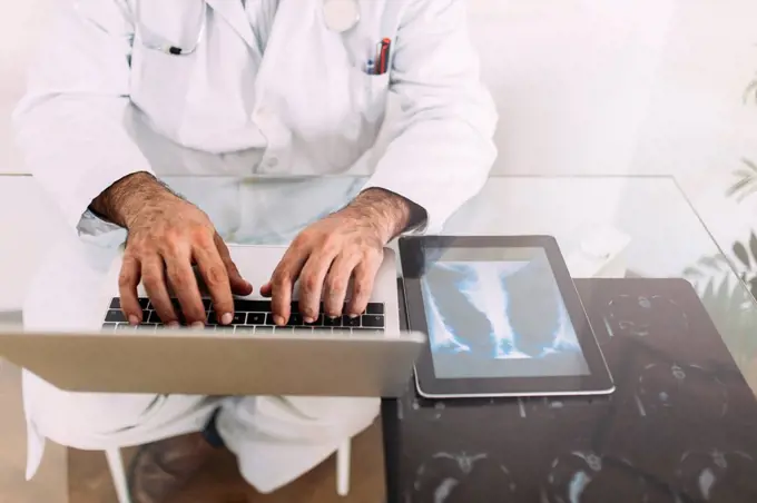 Senior male doctor working on laptop at desk in clinic