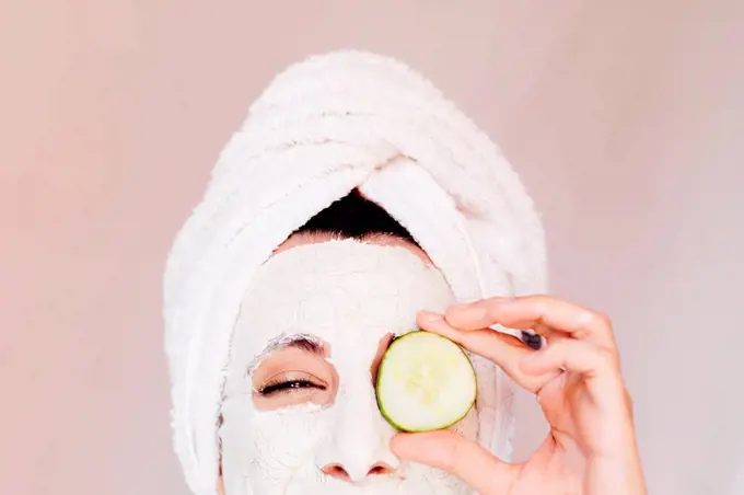 Young woman with beauty face mask covering eye with cucumber slice