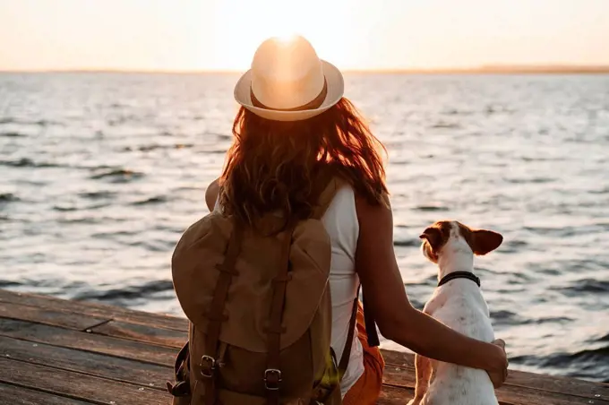 Woman with backpack and hat sitting by dog on pier