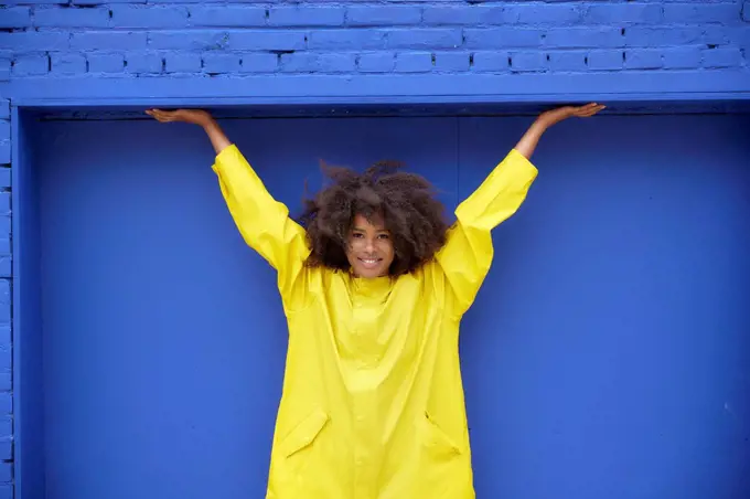 Smiling young Afro woman with arms raised in front of blue wall