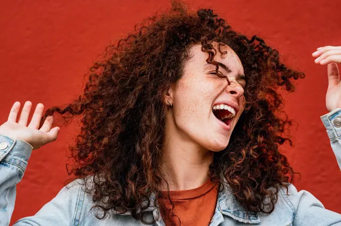 Cheerful young woman with curly hair singing in front of red wall