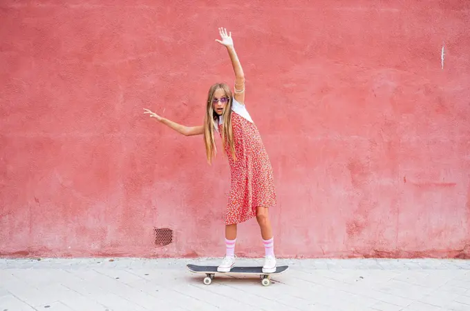 Pre-adolescent girl with hand raised skateboarding on footpath