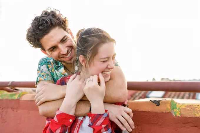 Young man embracing cheerful girlfriend on rooftop during weekend