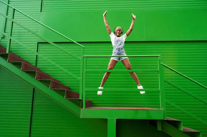 Happy woman with hands raised jumping while listening music on staircase in front of green wall